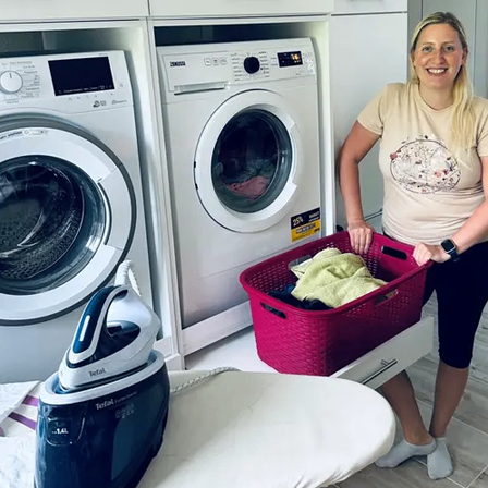 Eine Frau steht neben einem Waschraum und hält einen Korb mit Wäsche. Hinter ihr befinden sich zwei Waschmaschinen und ein Bügeleisen auf einem Bügelbrett.