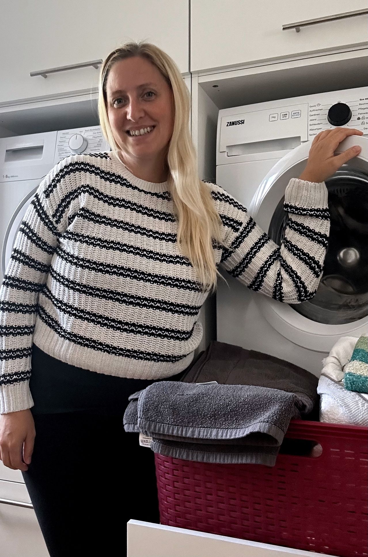 Eine Frau mit blonden Haaren steht in einer Waschküche und lehnt sich gegen eine Waschmaschine. Sie trägt einen schwarz-weiß gestreiften Pullover.