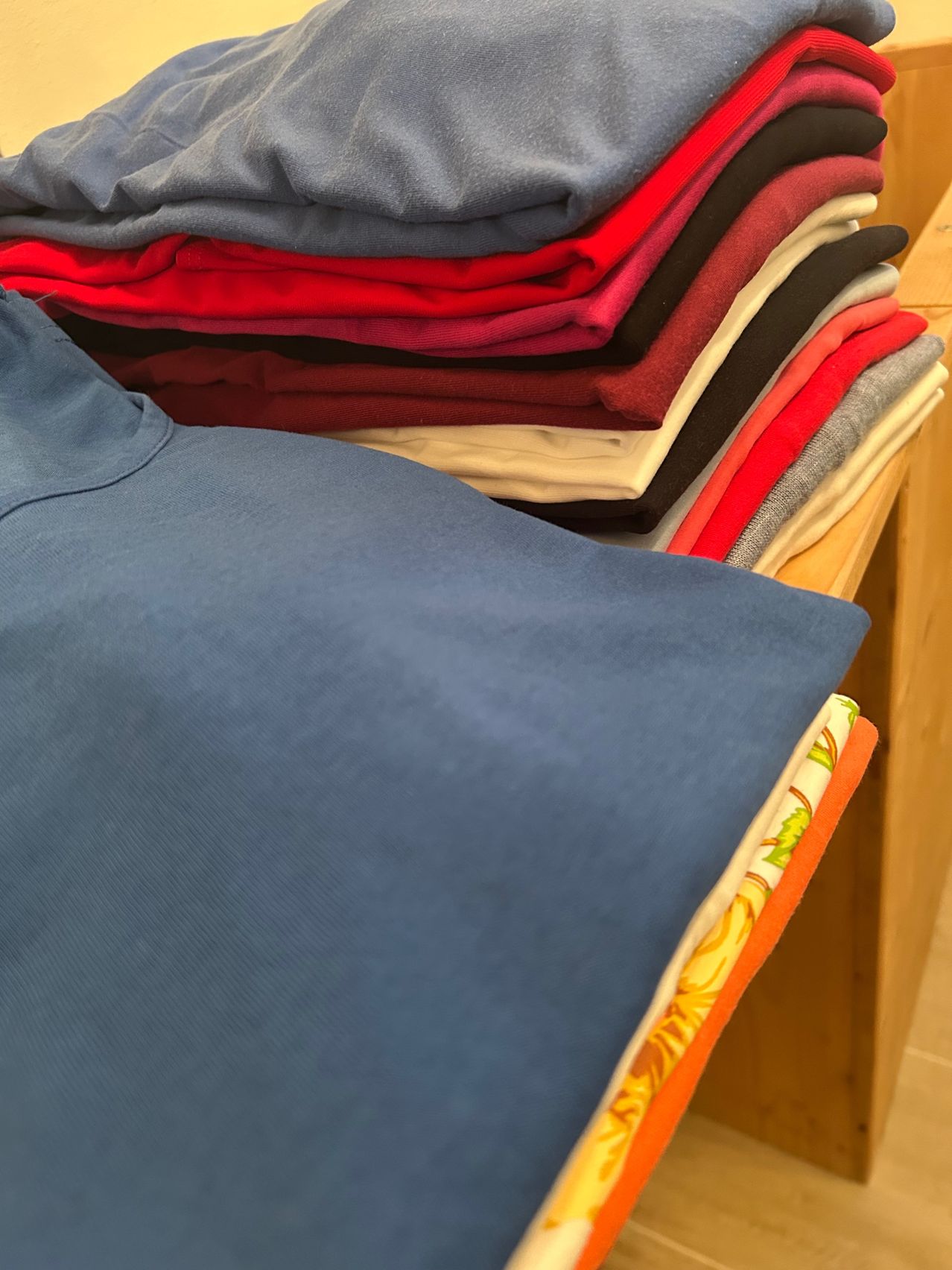 Ein Holzregal zeigt ordentlich gefaltete Hemden in verschiedenen Farben, darunter Blau, Rot, Schwarz und Grau. Das blaue Hemd ist vorne gefaltet.