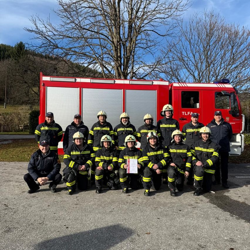 Eine Gruppe Feuerwehrleute in voller Uniform posiert für ein Foto vor einem roten Feuerwehrauto mit Bäumen im Hintergrund.