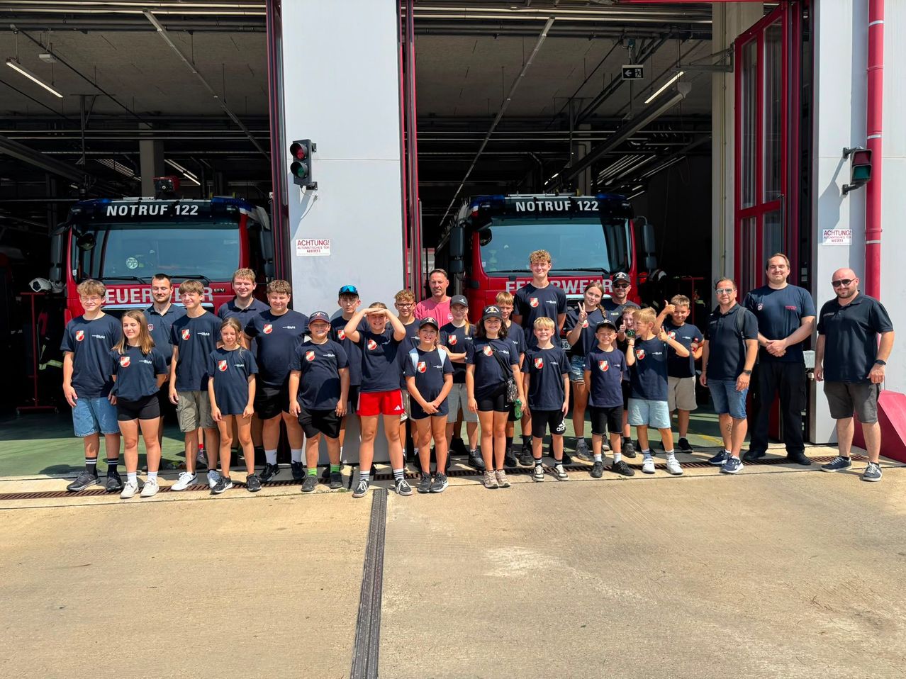 Eine Gruppe von Kindern und Erwachsenen posiert für ein Foto vor einer Feuerwache mit zwei roten Feuerwehrwagen.