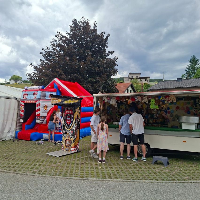 Menschen sind auf einem Jahrmarkt mit einem Hüpfburg und einem Stand mit Kuscheltieren. Ein Baum und Häuser sind im Hintergrund.