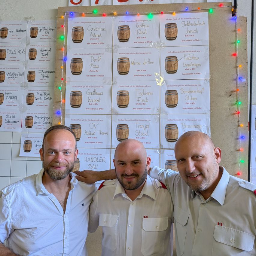 Drei Männer in weißen Uniformen stehen zusammen. Dahinter ist eine Wand mit Fässern und bunten Lichtern. Die Fässer haben Namen und Nachrichten darauf geschrieben.