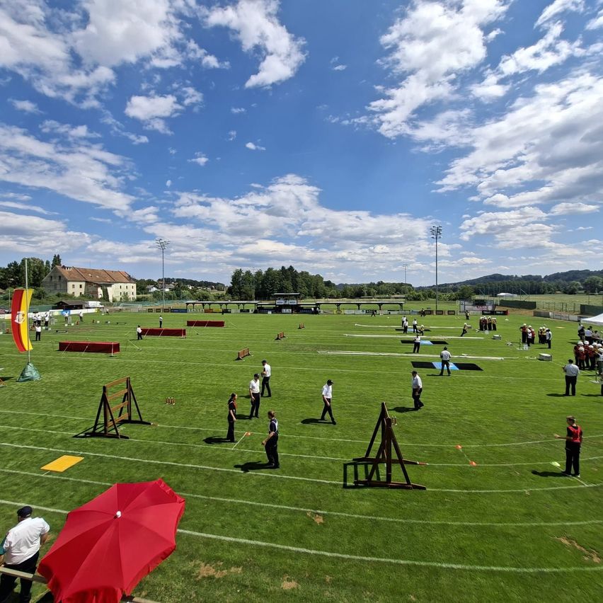A sports field with various obstacles and people in white uniforms. A red umbrella is on the left, and a flag is on the field.