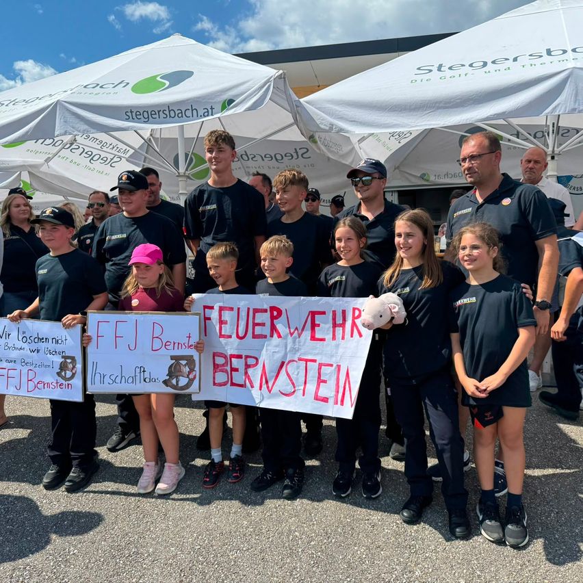 A group of children and adults are standing under white tents, holding signs that say FFJ Bernstein and Feuerwehr Bernstein. They are wearing matching black uniforms.