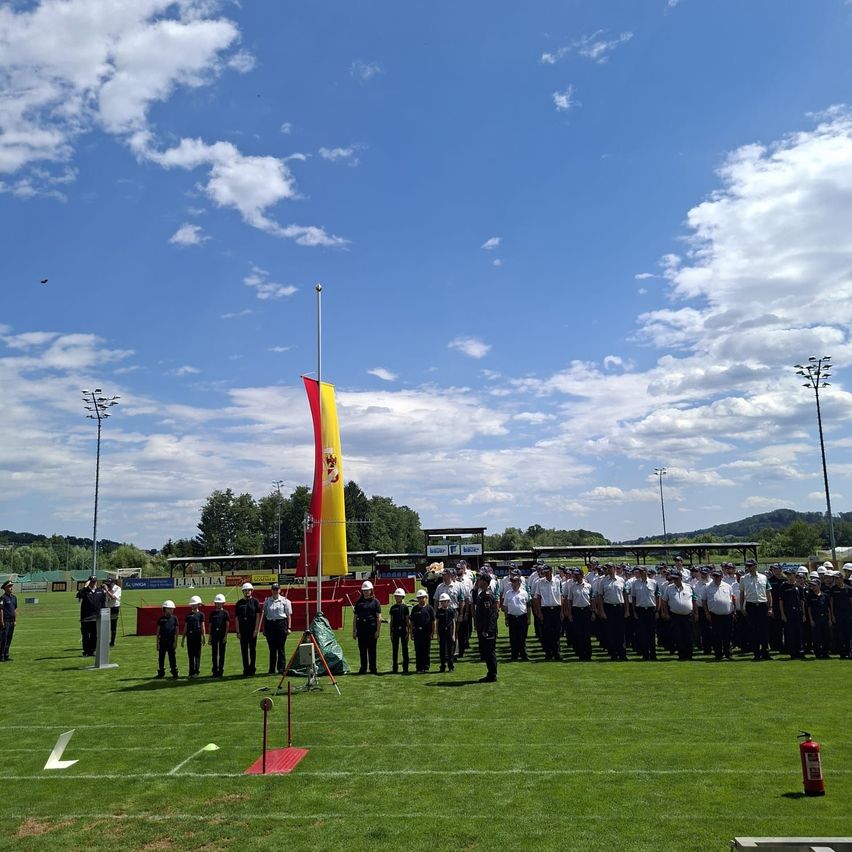 A group of uniformed individuals stands on a field under a blue sky with clouds, near a flagpole with a red and yellow flag.