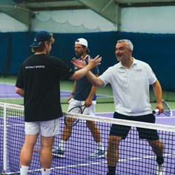 Drei Männer auf einem Tennisplatz. Einer schüttelt dem anderen die Hand, beide tragen Tennisausrüstung. Der dritte Mann steht hinter ihnen.