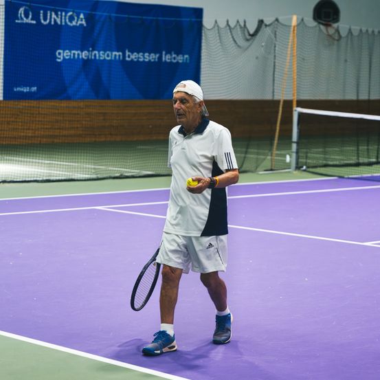 Ein älterer Mann geht auf einem Tennisplatz, hält einen Tennisball und einen Schläger in der Hand und trägt eine Kappe und Turnschuhe. Hinter ihm befindet sich ein blauer Banner mit dem Text 'Gemeinsam besser leben'.