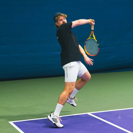 Ein Mann spielt Tennis auf einem Platz. Er trägt ein schwarzes T-Shirt, weiße Shorts und Turnschuhe. Er hält einen Tennisschläger und scheint mitten im Schwung zu sein.