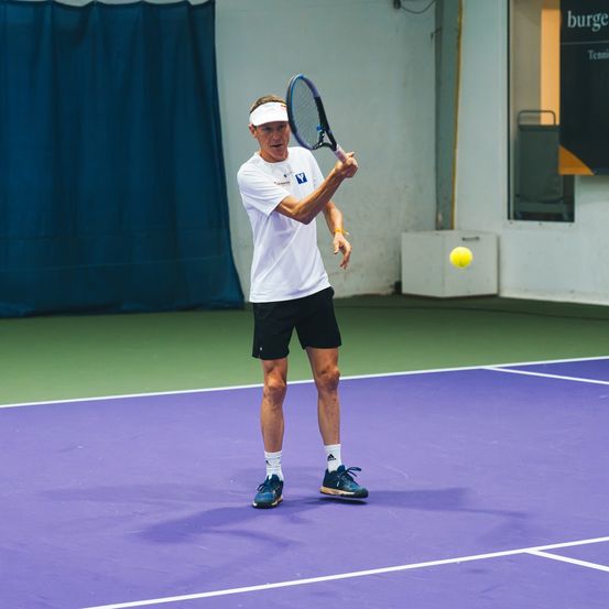 Ein älterer Mann spielt Tennis auf einem Platz, hält einen Schläger und bereitet sich darauf vor, einen gelben Ball zu schlagen.
