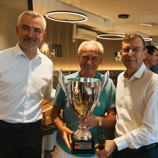 Drei Männer stehen in einem Innenbereich und lächeln. Der Mann in der Mitte hält einen Pokal, und der Mann auf der rechten Seite hält ein Mikrofon. Der Mann auf der linken Seite trägt einen Gürtel.