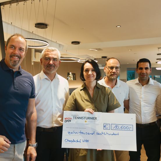 Fünf Personen stehen in einem Innenraum zusammen und halten einen Scheck mit Text und Zahlen in blauer Tinte. Die Frau in der Mitte lächelt in die Kamera.