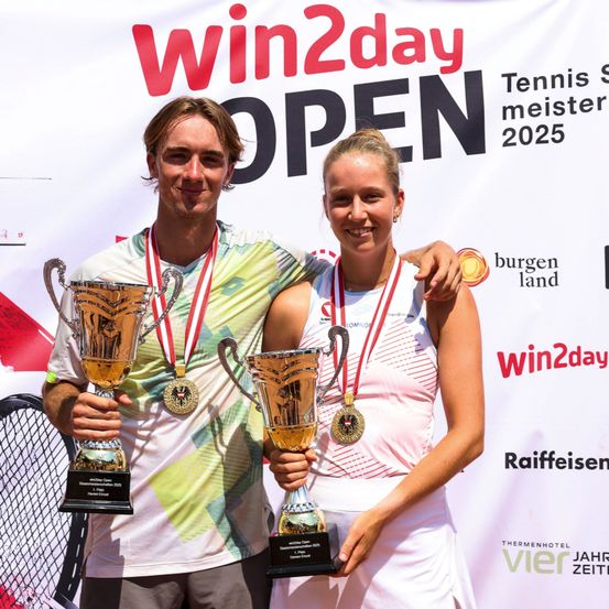 Ein Mann und eine Frau, beide mit Medaillen, stehen zusammen und halten Pokale bei einem Tennis-Event. Das Banner hinter ihnen sagt 'Win2day' und 'Tennis Meister 2025'.
