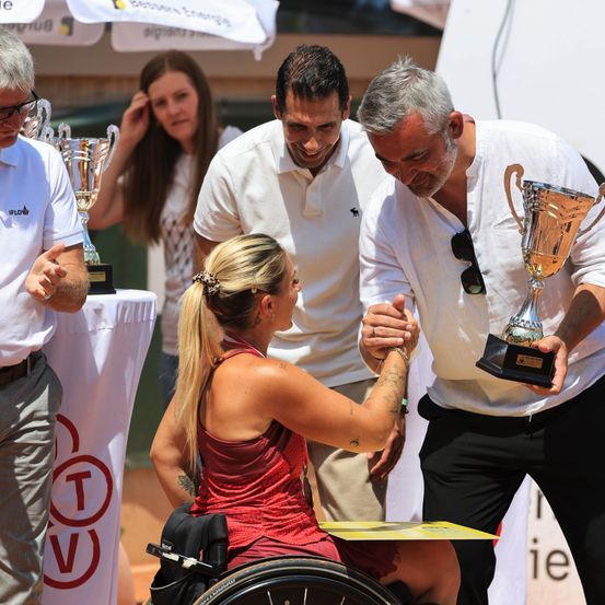 Eine Frau im Rollstuhl hält die Hand eines Mannes, der einen Pokal hält. Zwei Männer stehen hinter ihnen, und ein weiterer Mann mit einem Pokal ist in der Nähe. Eine Frau blickt aus dem Hintergrund.