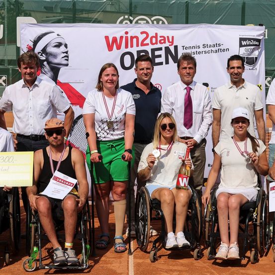 Eine Gruppe von Tennisspielern in Rollstühlen posiert für ein Foto mit Offiziellen auf einem Tennisplatz. Sie halten Medaillen und eine Flasche Wein. Ein Banner mit Win2day und OPEN ist hinter ihnen.