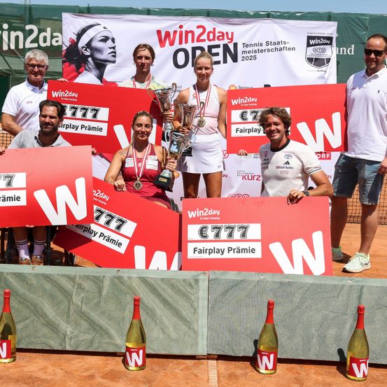 Tennisspieler stehen auf einem Tennisplatz, halten Pokale und heben große rote Plakate mit €777 hoch. Ein Mann und eine Frau halten ein Schild mit der Aufschrift 'Win2Day OPEN', während andere für die Kamera posieren. Flaschen Wein sind vor den Spielern ausgestellt.