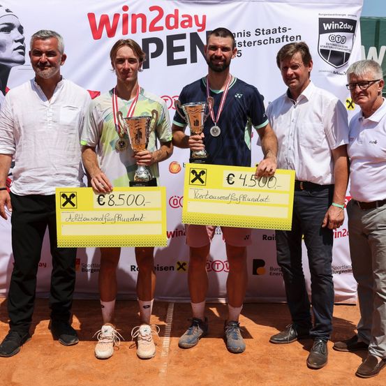 Fünf Männer stehen auf einem Tennisplatz, halten Pokale und einen gelben Scheck. Zwei Männer tragen Medaillen, und zwei halten Pokale. Dahinter befindet sich ein Banner mit Text und Logo.