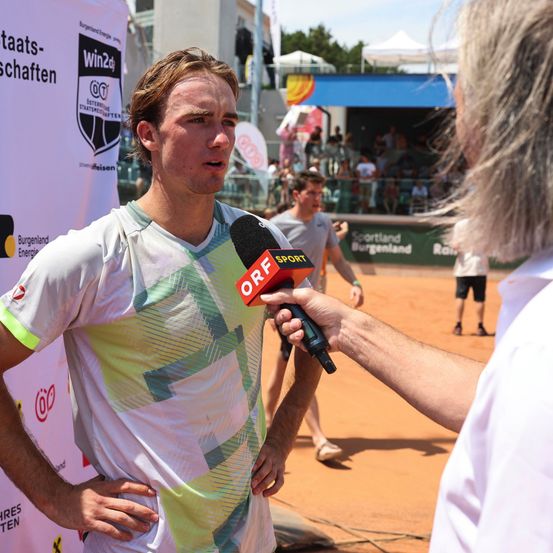 Ein Mann in einem grün-weißen Shirt wird von einem Reporter interviewt, der ein Mikrofon hält. Sie stehen auf einem Tennisplatz mit einem Banner und einer Menge im Hintergrund.