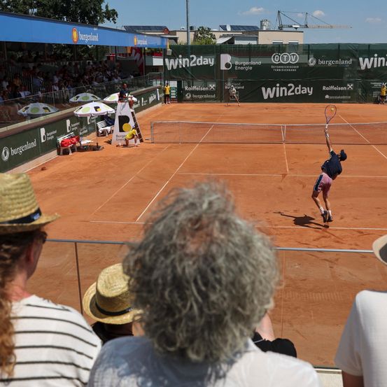 Ein Tennismatch findet auf einem Sandplatz statt. Zuschauer sitzen und beobachten die Spieler. Ein Spieler springt, um den Ball zu schlagen. Werbung ist an den Platzbarrieren sichtbar.