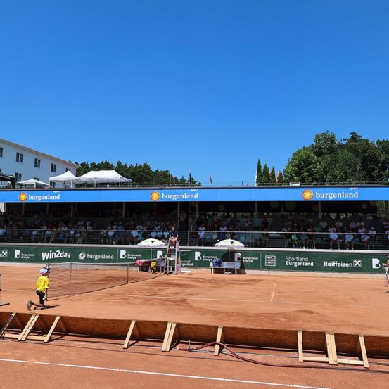 Ein Tennisplatz im Freien mit einem blauen Banner, das Burgenland bewirbt, Zuschauer auf den Tribünen, ein Kind mit Tennisschläger und eine Person, die auf dem Platz kniet.