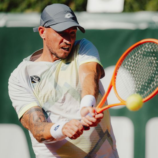 Ein männlicher Tennisspieler mit Tattoos am Arm schwingt einen orangefarbenen Schläger, um einen gelben Ball zu schlagen.