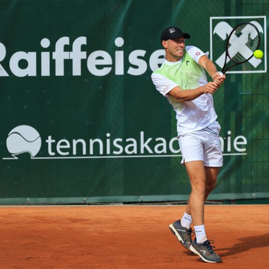 Ein Tennisspieler schlägt einen Ball auf einem Tennisplatz mit einer Sandfläche. Er trägt ein grün-weißes Shirt, weiße Shorts und graue Turnschuhe.