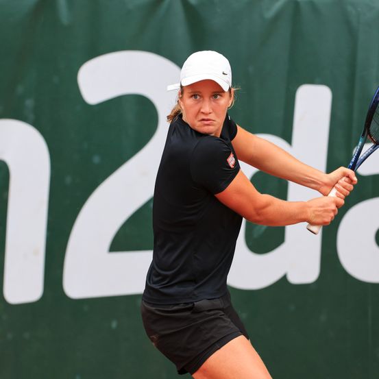 Eine Tennisspielerin schwingt einen Schläger bei einem Match, trägt ein schwarzes Shirt, schwarze Shorts und eine weiße Kappe. Hinter ihr ist eine grüne Kulisse mit weißen Buchstaben.