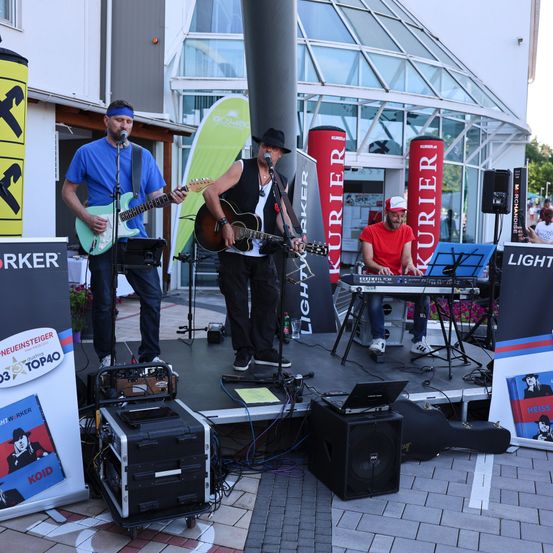 Drei Musiker spielen auf einer Bühne in einem Außenbereich. Einer spielt Gitarre, ein anderer spielt Klavier und der dritte spielt ebenfalls Gitarre. Im Hintergrund befinden sich Lautsprecher, Banner und ein Gebäude.