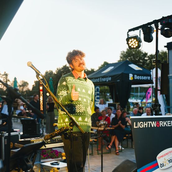 Ein Mann in einem grünen Pullover steht auf der Bühne mit einem Mikrofon und gibt einen Daumen hoch. Hinter ihm sitzt eine Menschenmenge vor einem Zelt mit dem Wort Bessere darauf. Bühnenlichter sind an einem Ständer montiert.