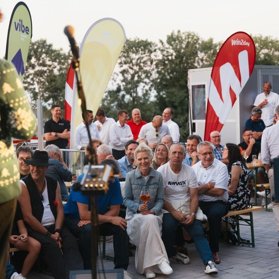 Eine Outdoor-Versammlung mit Menschen, die auf Bänken sitzen und stehen. Eine Frau hält ein Weinglas, während andere zusehen. Ein Mann steht hinter einem Mikrofon, und Banner sind im Hintergrund sichtbar.