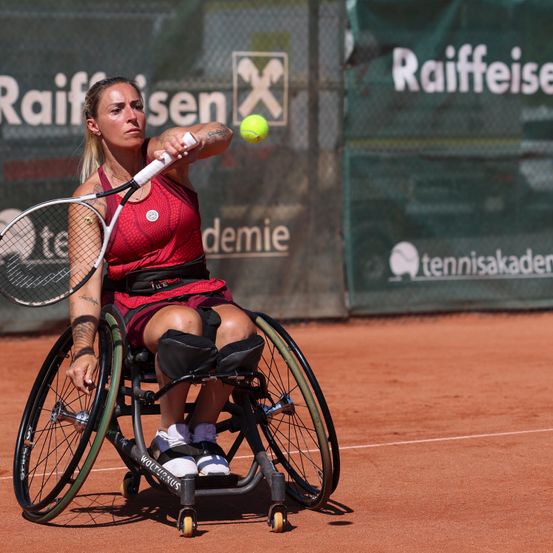 Eine Tennisspielerin im Rollstuhl, in einem roten Outfit, ist dabei, einen Tennisball auf einem Sandplatz zu schlagen. Der Platz ist von einem Zaun mit Sponsorenwerbung umgeben.