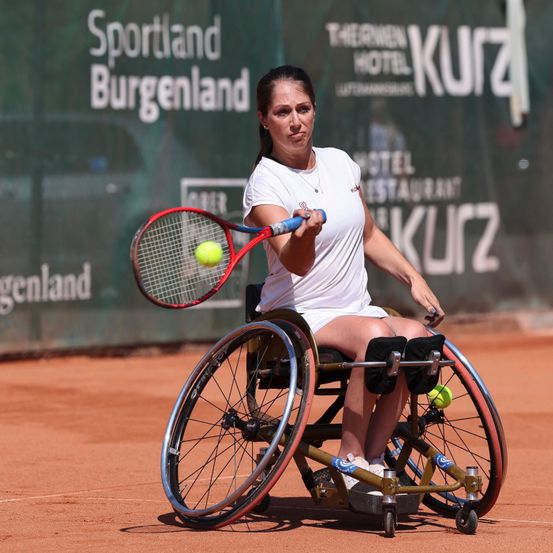 Eine Frau im Rollstuhl spielt Tennis auf einem Sandplatz. Sie hält einen Tennisschläger und schlägt einen Tennisball. Der Platz ist von einer grünen Mauer mit Werbung umgeben.