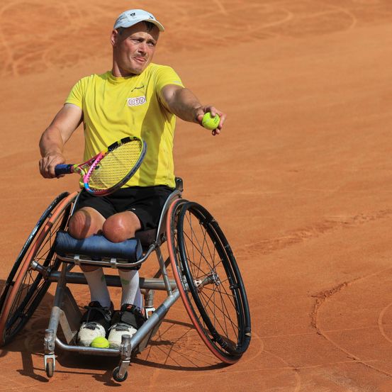 Ein Mann in einem gelben T-Shirt sitzt auf einem Rollstuhl auf einem Tennisplatz und hält eine Tennisschläger und einen Tennisball.