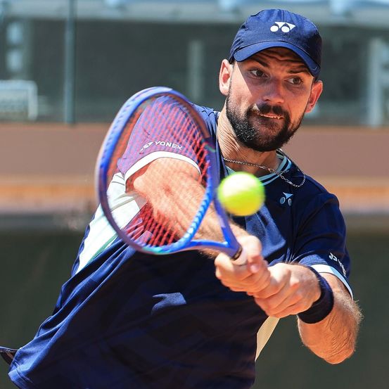 Ein männlicher Tennisspieler in einem blauen Shirt schlägt einen Tennisball mit seinem Schläger.