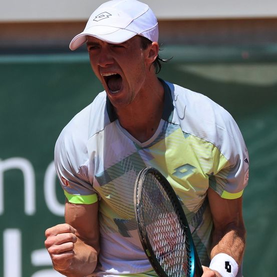 Ein Tennisspieler schreit im Triumph, hält einen Tennisschläger und trägt ein weißes Shirt mit geometrischem Muster.