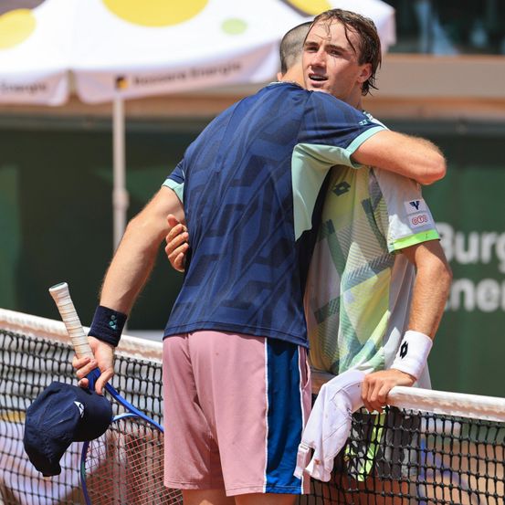 Zwei Tennisspieler umarmen sich auf einem Platz mit einem blau-weißen Netz und einem weißen Schirm im Hintergrund.