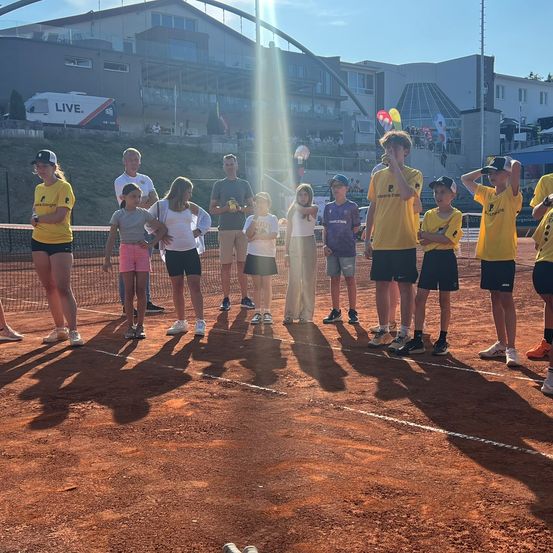 Eine Gruppe von Menschen in gelben Shirts und Shorts steht auf einem Tennisplatz, einige halten Tennisschläger und andere beobachten. Im Hintergrund befinden sich Gebäude mit Glasfenstern und ein Schild mit der Aufschrift 'LIVE'.