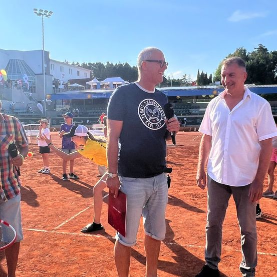 Zwei Männer stehen auf einem Tennisplatz mit einem Mikrofon. Einer trägt ein blaues T-Shirt mit Logo. Ein Kind übt Tennis in der Nähe. Zuschauer und Gebäude sind im Hintergrund.