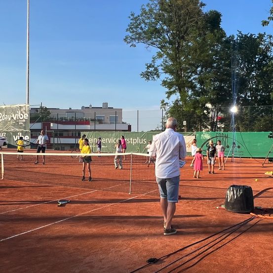 Tennisspieler auf einem Platz mit Netz, ein Mann steht dahinter und eine Fahne im Hintergrund.