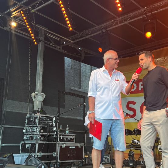 Zwei Männer stehen auf einer Bühne mit Musikequipment im Hintergrund. Ein Mann spricht in ein Mikrofon, während der andere zuhört.