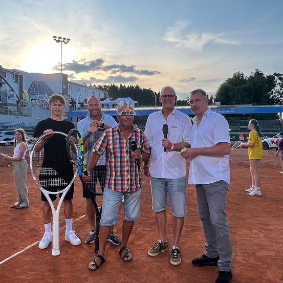 Fünf Männer stehen auf einem Tennisplatz und halten Tennisschläger. Sie sind von Zuschauern umgeben und in einer Umgebung mit blauem Himmel und Wolken.