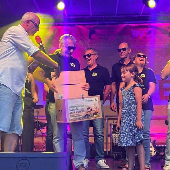 Eine Gruppe von Menschen auf einer Bühne, ein Mann hält ein Mikrofon, ein anderer ein Kasten, und ein junges Mädchen vor ihnen. Dahinter ein Banner mit dem Text 'BVZ'. Die Umgebung ist lebhaft mit Lichtern und Musikinstrumenten.