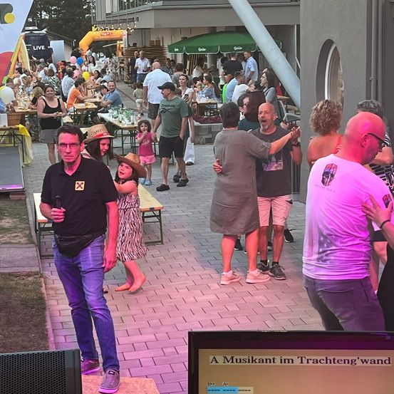 Menschen versammeln sich in einem Außenbereich, einige sitzen an Tischen, andere tanzen. Ein Mann mit Mikrofon steht auf einer Bühne, und andere interagieren in der Nähe. Bunte Luftballons und ein grüner Regenschirm sind sichtbar.