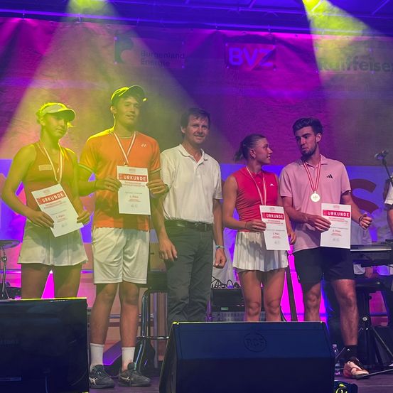 Fünf Personen stehen auf einer Bühne und halten Urkunden und Medaillen in der Hand. Im Hintergrund ist ein Plakat mit Burgenland Energie und BVZ zu sehen. Auf der Bühne sind Musikinstrumente und Lautsprecher aufgebaut.