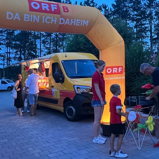 Menschen versammeln sich um einen gelben Imbisswagen unter einem orangefarbenen Bogen. Eine Frau und ein Kind interagieren mit einem Ballonkünstler. Dahinter arbeitet ein Mann am Wagen.