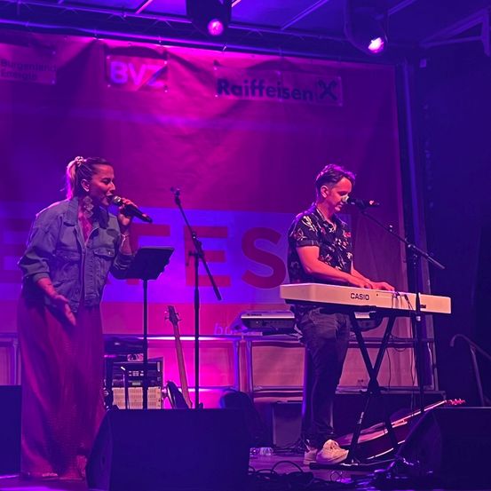 Ein Mann und eine Frau treten auf der Bühne mit lila Beleuchtung auf. Die Frau hält ein Mikrofon, und der Mann spielt eine Tastatur. Ein Banner mit dem Text RaiffeisenX steht hinter ihnen.