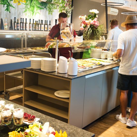 Ein Restaurantbuffet mit einem Mann, der Essen serviert. Ein Mann in einem Hut und ein anderer in einem weißen T-Shirt warten. Dahinter steht ein weiterer Mann. Teller, Blumen und Flaschen sind auf der Theke.