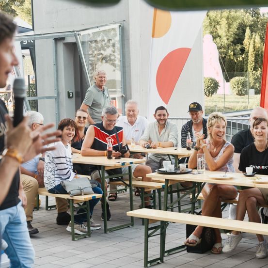 Eine Gruppe von Menschen sitzt draußen an Tischen, lächelt und lacht. Ein Mann steht vor ihnen mit einem Mikrofon. Die Tische haben Teller, Tassen und Taschen. Dahinter befindet sich eine Wand mit einer Tür und einer roten Flagge.