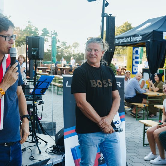 Zwei Männer stehen auf einer Bühne mit Lautsprechern und einem Mikrofon. Ein Mann trägt ein rot-weiß-blaues Shirt und eine Brille. Der andere Mann trägt ein schwarzes Shirt mit dem Wort Boss darauf. Dahinter sind Tische und Stühle mit sitzenden Leuten. Es gibt Zelte und Bäume in der Ferne.