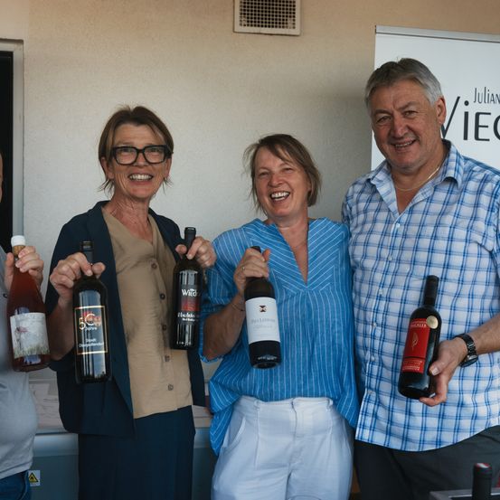 Vier Personen stehen zusammen und halten Weinflaschen. Die beiden Frauen lächeln in die Kamera. Der Mann rechts trägt ein kariertes Hemd und der Mann links trägt ein weißes Hemd.